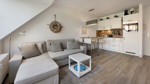 A modern living room with a sectional sofa, a small white table, a kitchen area with white cabinets, and a dining table with chairs.