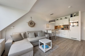 A modern living room with a sectional sofa, a small white table, a kitchen area with white cabinets, and a dining table with chairs. - Holiday Apartment 'Wohnanlage Am Mühlenteich' with Balcony and Wi-Fi (Timmendorfer Strand)