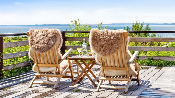 Zwei Holzstühle mit Fellpolsterung blicken auf eine Holzterrasse mit Blick auf ein Gewässer.