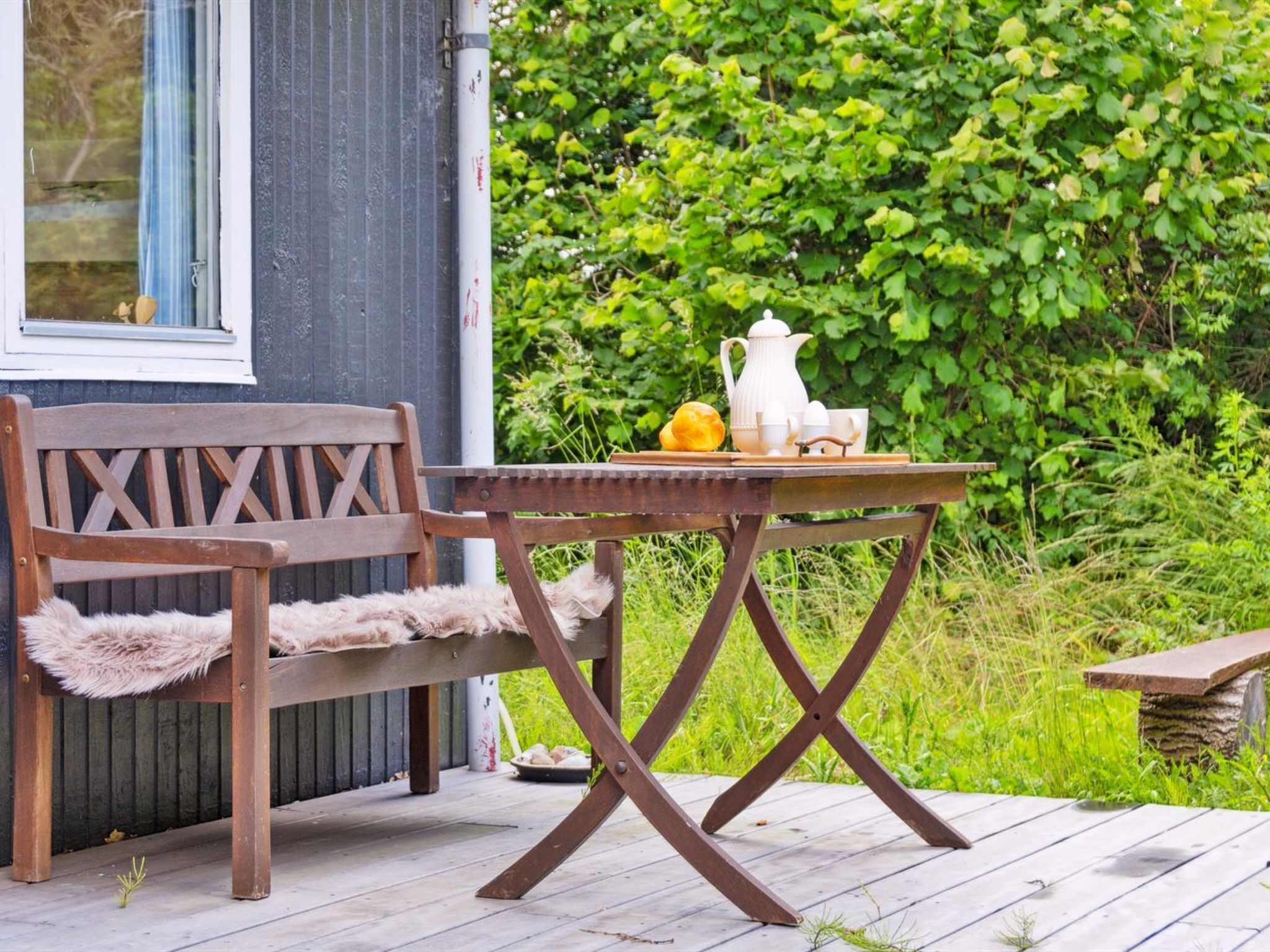 A wooden bench with a fur throw, a table with a white pitcher, cups, and a bowl of fruit, set on a wooden deck with a garden wall in the background.