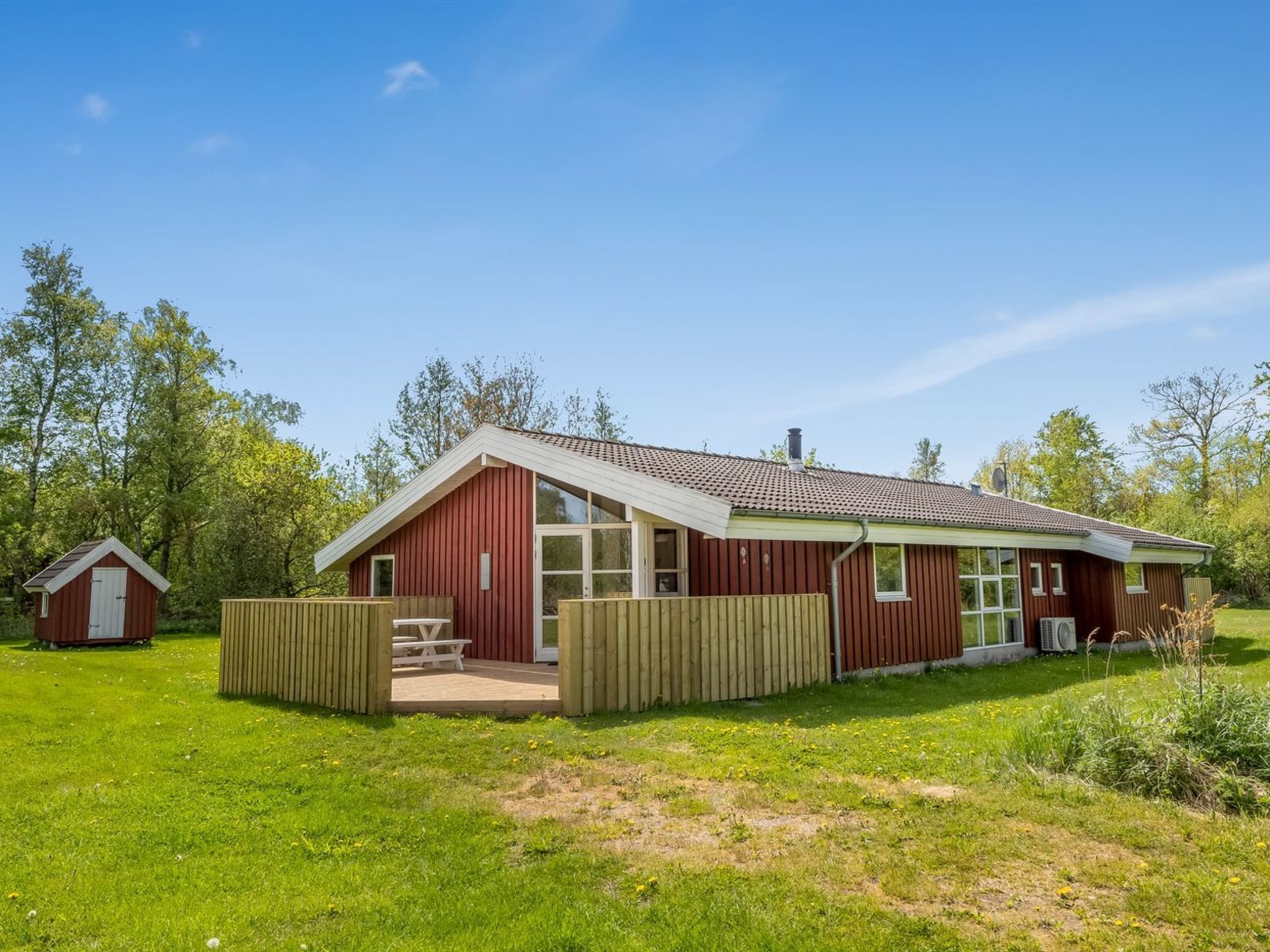 A red wooden house with a large window, a small attached shed, and a picnic table in the yard.