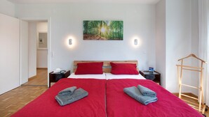 Una cama doble con ropa de cama roja, un cabecero de madera, una mesita de noche con una lámpara y un cuadro de un bosque en la pared.