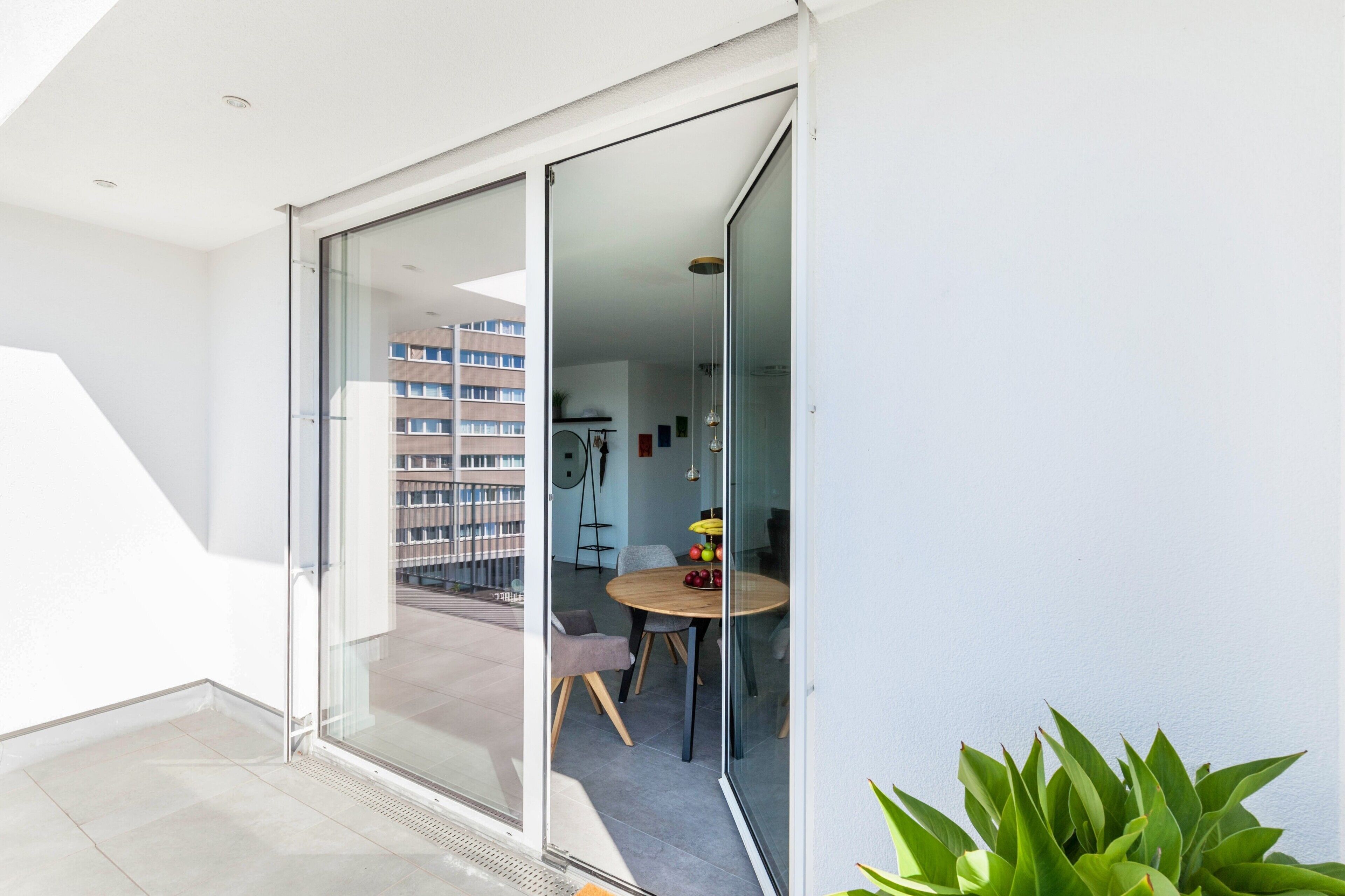 Un appartement moderne doté d’un balcon, d’un coin repas avec une table ronde et des chaises, et offrant une vue sur un immeuble en arrière-plan.