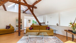 Un salon spacieux avec un plafond haut, des poutres apparentes, une table basse en verre et un canapé agrémenté de coussins colorés.