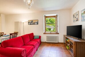 A living room with a red sofa, a television on a wooden stand, a dining table with chairs, a radiator, and a window with a view of greenery. - La Madonnina Apartment for 2 in Tenno (Tenno)