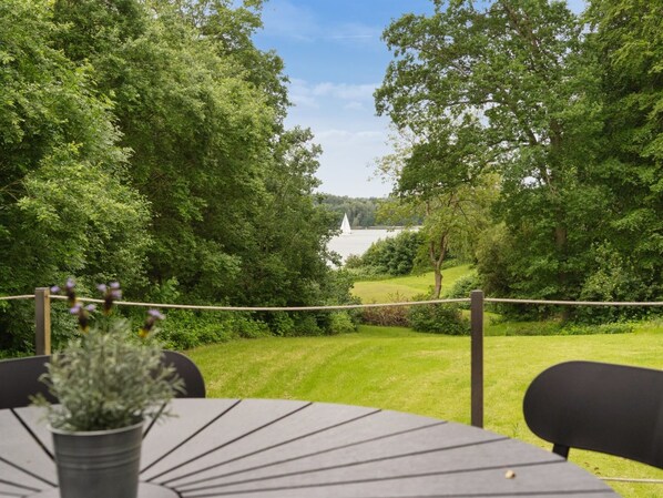 A table with chairs and a potted plant, overlooking a grassy area with trees and a body of water.