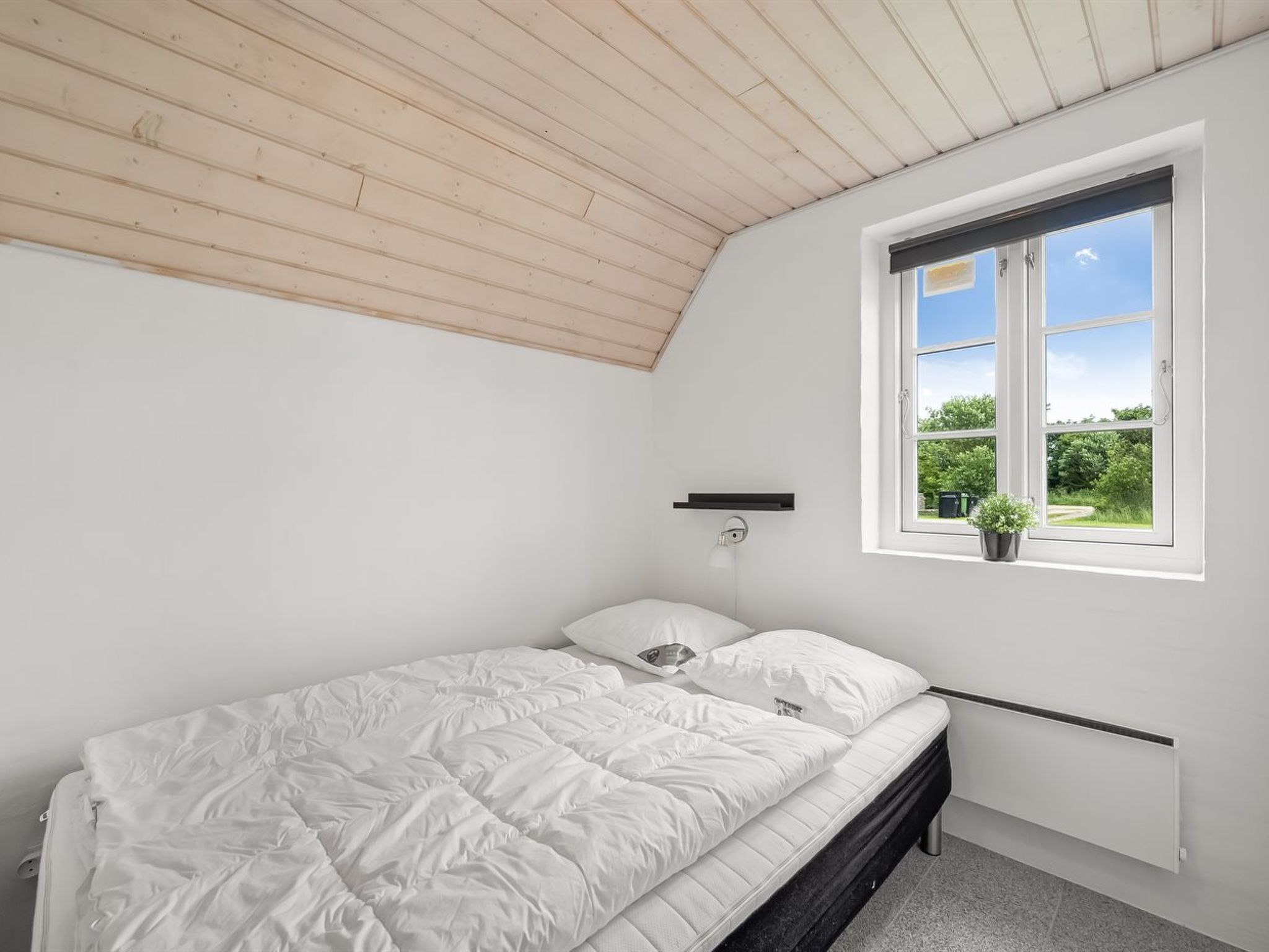 A bedroom with a sloped ceiling, a single bed, a bedside table, a window with a view of greenery, and a small potted plant.