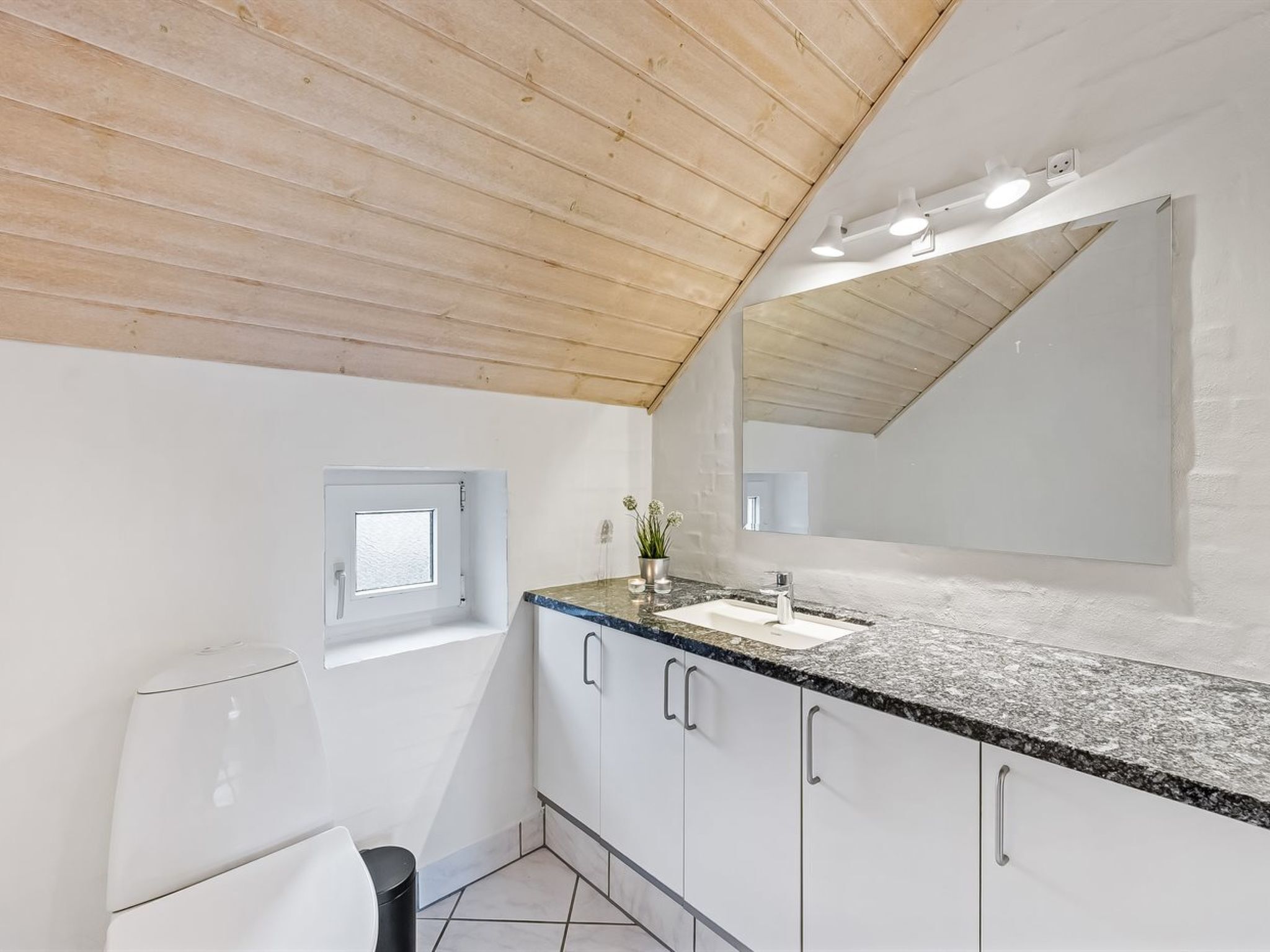 Ein modernes Badezimmer mit schrägverdeckelter Decke, einem großen Spiegel, einer Granit-Arbeitsplatte und weißen Schränken.