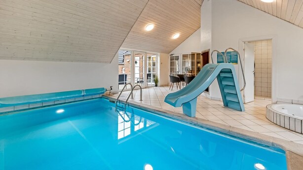 An indoor swimming pool with a slide, a hot tub, and a seating area with a view of the outside.