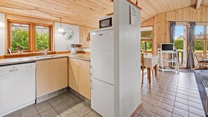A modern kitchen with wooden cabinets, a built-in dishwasher, and a large refrigerator. There is a dining area with a table and chairs, and a view of the outdoors through large windows. - "Åsa" - 500m to the fjord by Interhome (Tarm)