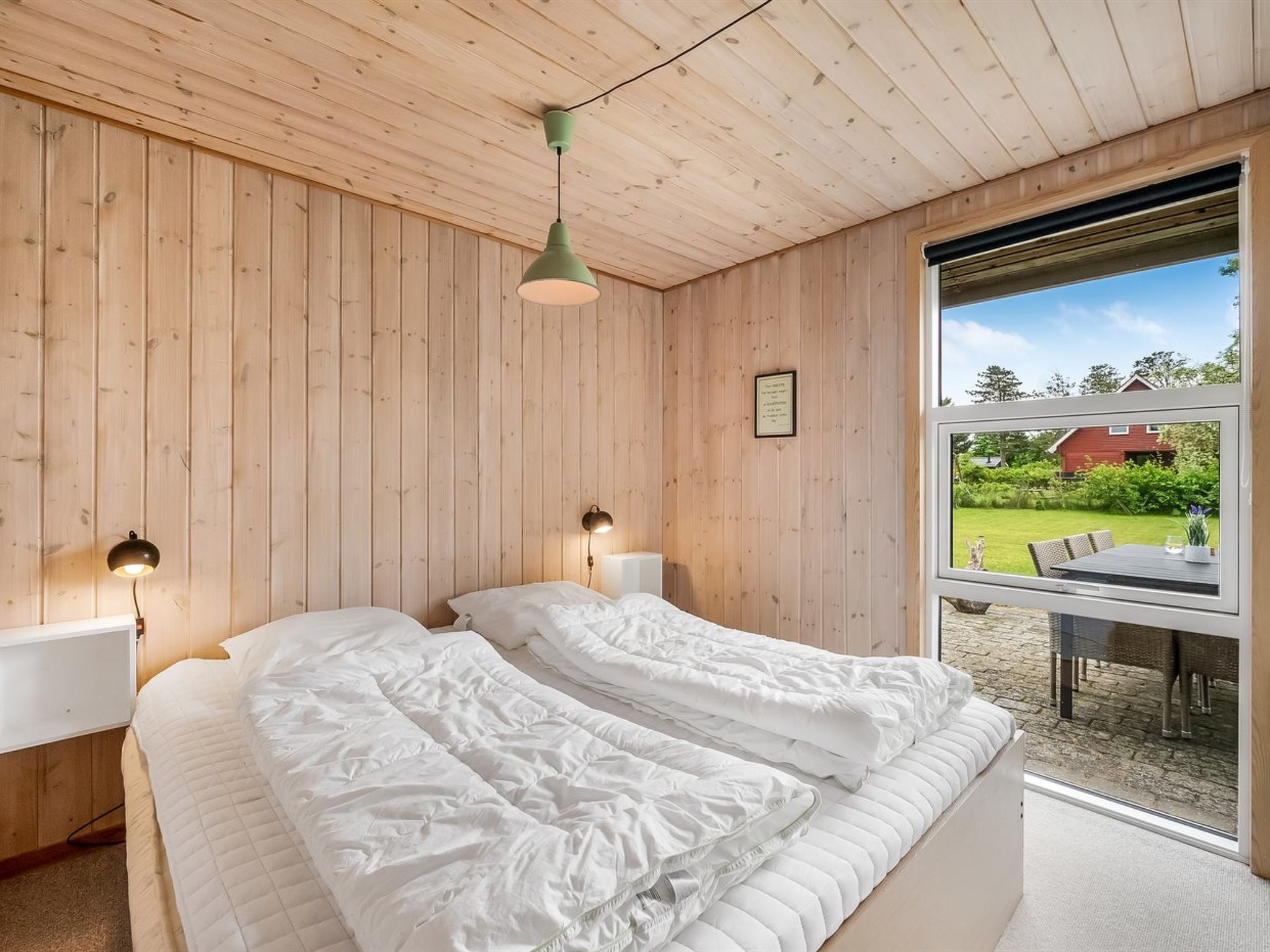 Ein Schlafzimmer mit Holzboden, einem Bett mit weißer Bettwäsche, zwei Nachttischlampen und einem Fenster mit Blick auf ein Haus und einen Garten.