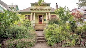 Une maison verte avec un porche à l’avant, des marches menant à la porte et un jardin où poussent différentes plantes et fleurs.