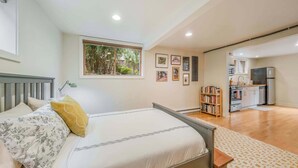 Une chambre comprenant un lit, une table de chevet, une fenêtre donnant sur un espace verdoyant, un coin cuisine équipé d’appareils électroménagers et une bibliothèque.