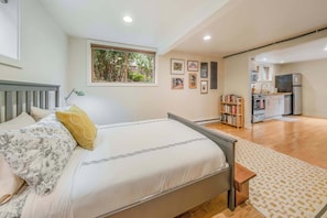 Un dormitorio con cama, mesita de noche, ventana con vistas a zonas verdes, kitchenette con electrodomésticos y una estantería.