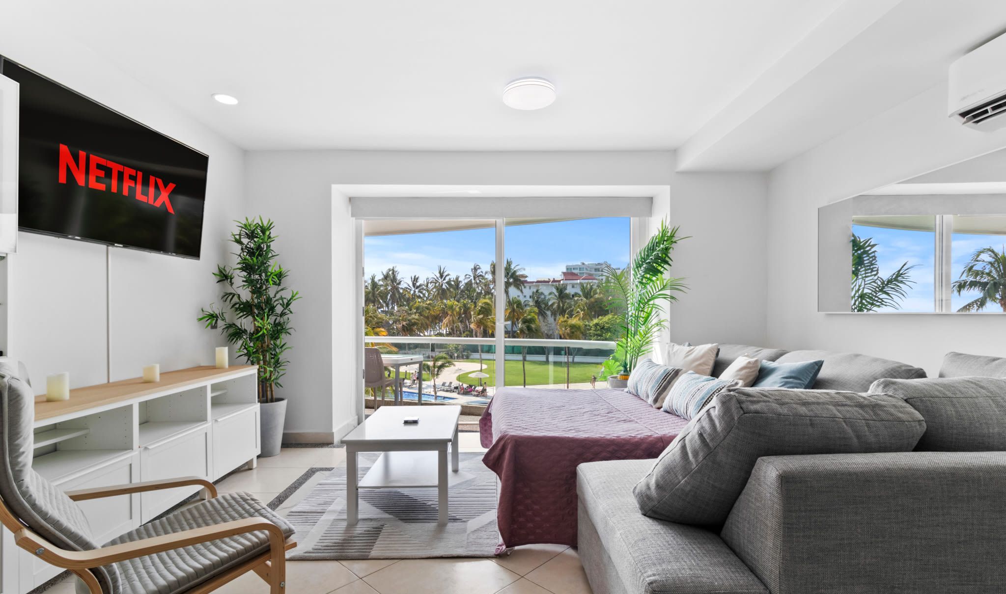 A modern living room with a flat-screen TV, a sofa, a coffee table, and a view of a pool and palm trees.