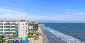 A beachfront hotel with multiple floors, a swimming pool, and a beach with umbrellas and lounge chairs.
