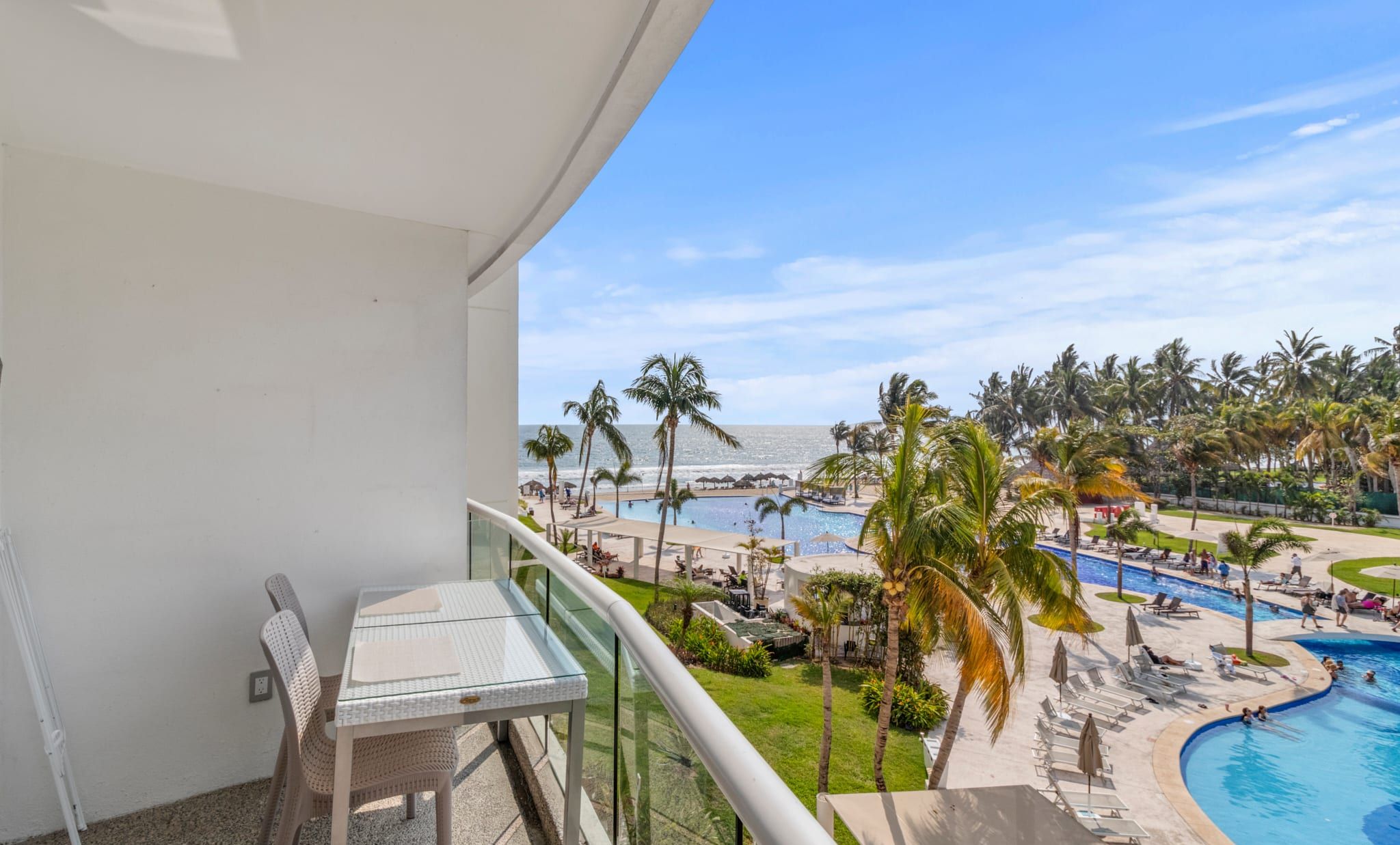 A balcony with a view of a resort pool area, including a swimming pool, lounge chairs, and palm trees.