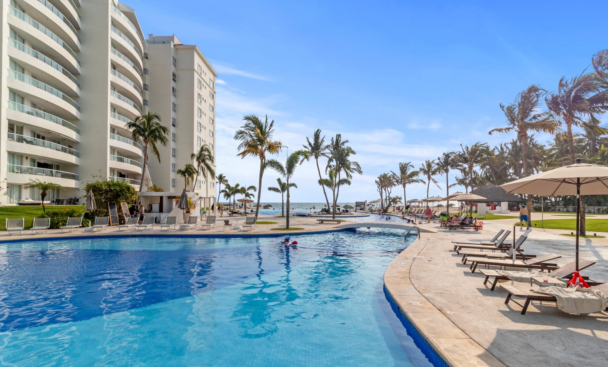 A resort pool area with lounge chairs and umbrellas, surrounded by palm trees and a large building.