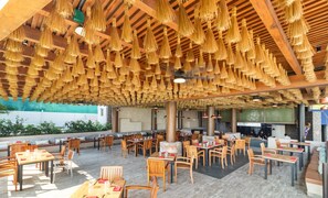 An outdoor dining area with wooden tables and chairs, a covered seating area with hanging tassels, and a view of the beach.