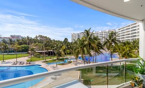 Un balcon d’hôtel donnant sur une piscine, des palmiers et un grand bâtiment en arrière-plan.