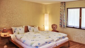 Un lit double avec une literie à motifs floraux, une table de chevet avec une lampe, une commode et une fenêtre avec des rideaux.