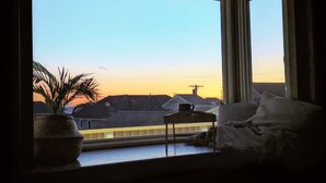 Une chambre dotée d’une grande fenêtre donnant sur un paysage côtier et offrant une vue sur le coucher du soleil, ainsi que d’un lit et d’une petite table sur laquelle est posée une plante.