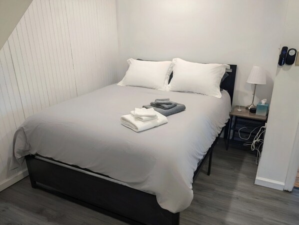 Un lit bien fait, avec du linge de lit blanc et gris, une table de chevet avec une lampe et un téléphone fixé au mur.