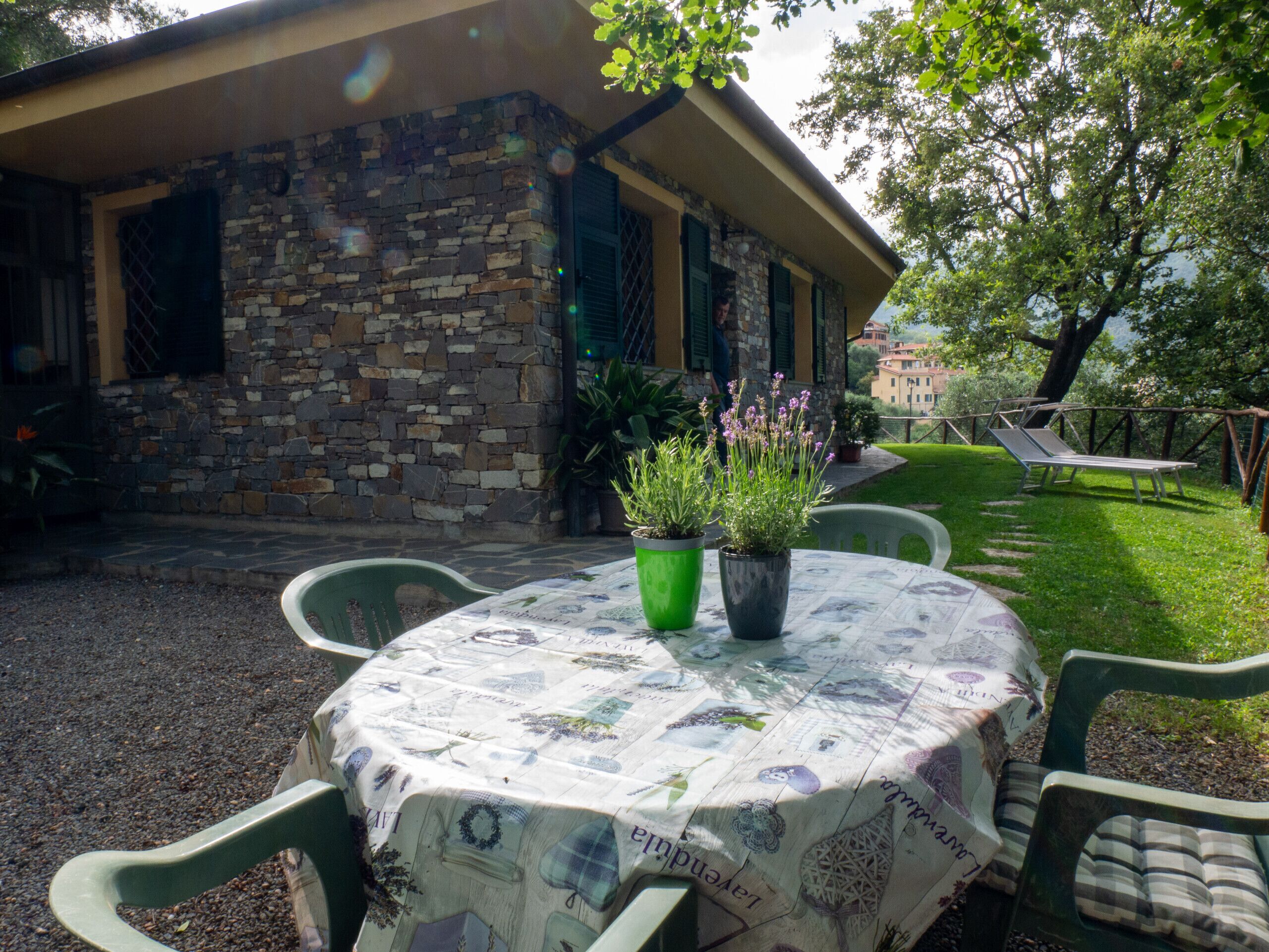 An outdoor seating area with a stone building, green chairs, a table with a floral tablecloth, and potted plants.