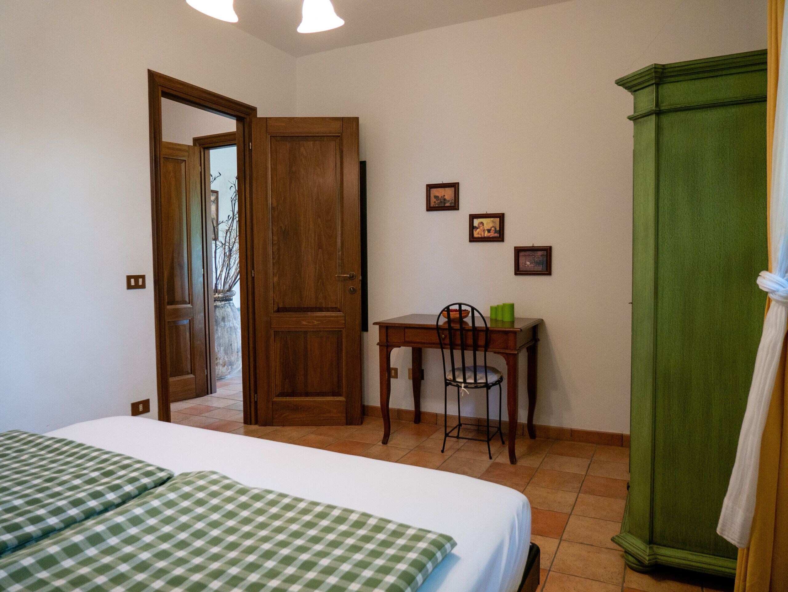 A bedroom with a bed, a desk, and a chair. There is a wardrobe and a door leading to another room.
