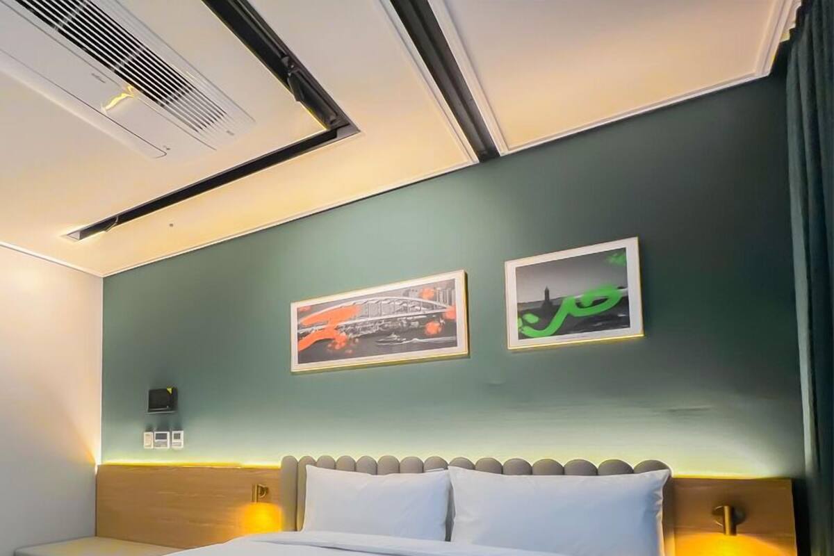 Habitación de hotel con cama, cabecera, dos lámparas fijadas a la pared y dos cuadros enmarcados.