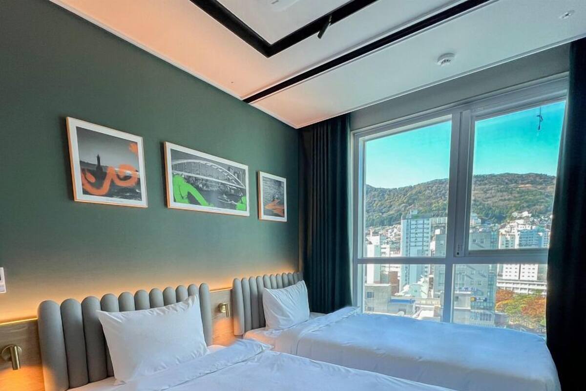 Habitación de hotel con dos camas, un ventanal grande con vistas a la ciudad y tres cuadros en la pared.