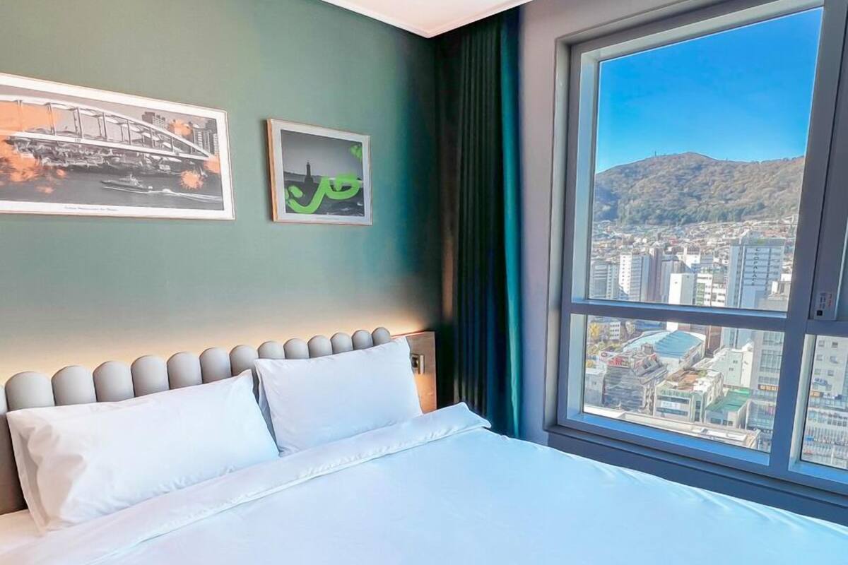 Habitación de hotel con un ventanal grande que ofrece vistas a la ciudad, una cama con sábanas blancas y dos cuadros en la pared.