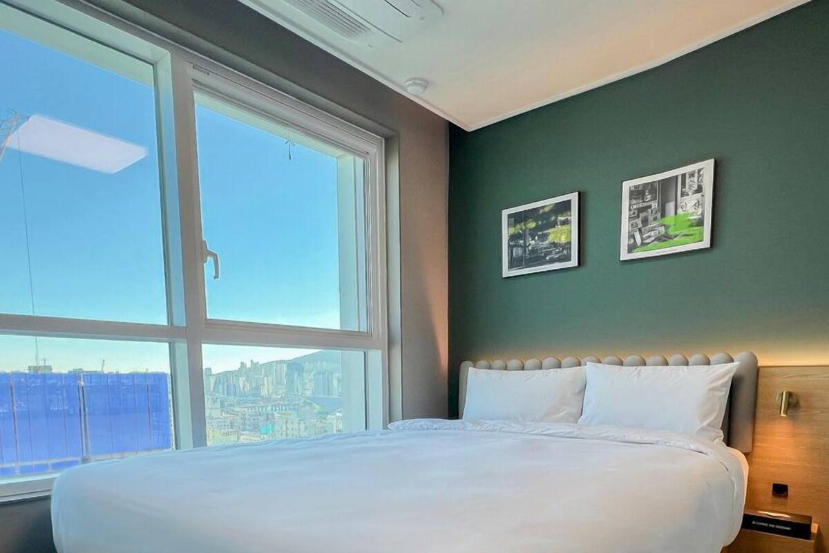 Habitación de hotel con una cama grande, vista a la ciudad a través de un ventanal amplio y dos cuadros en la pared.
