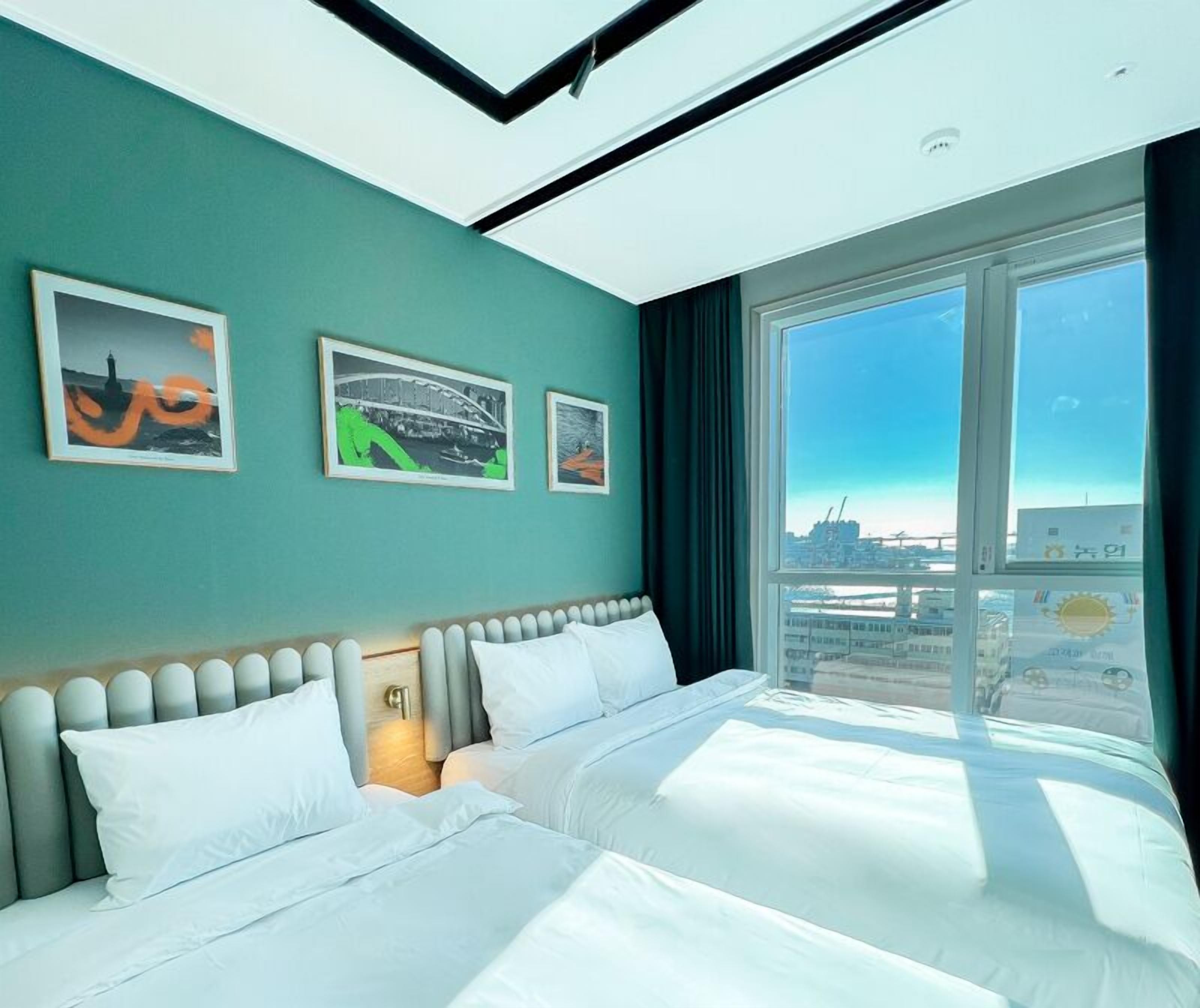 A modern hotel room with a large bed, two wall-mounted artworks, and a view of a cityscape through large windows.