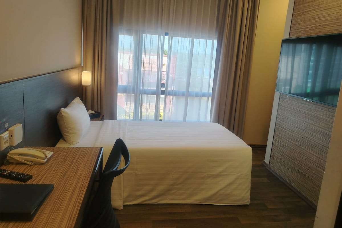 Quarto de hotel com cama, secretária com telefone e um caderno, uma cadeira e uma janela com cortinas.