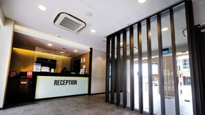 A reception area with a reception desk labeled 'RECEPTION', a glass door entrance, and a ceiling-mounted air conditioning unit. - The Corner House (Kuala Belait)