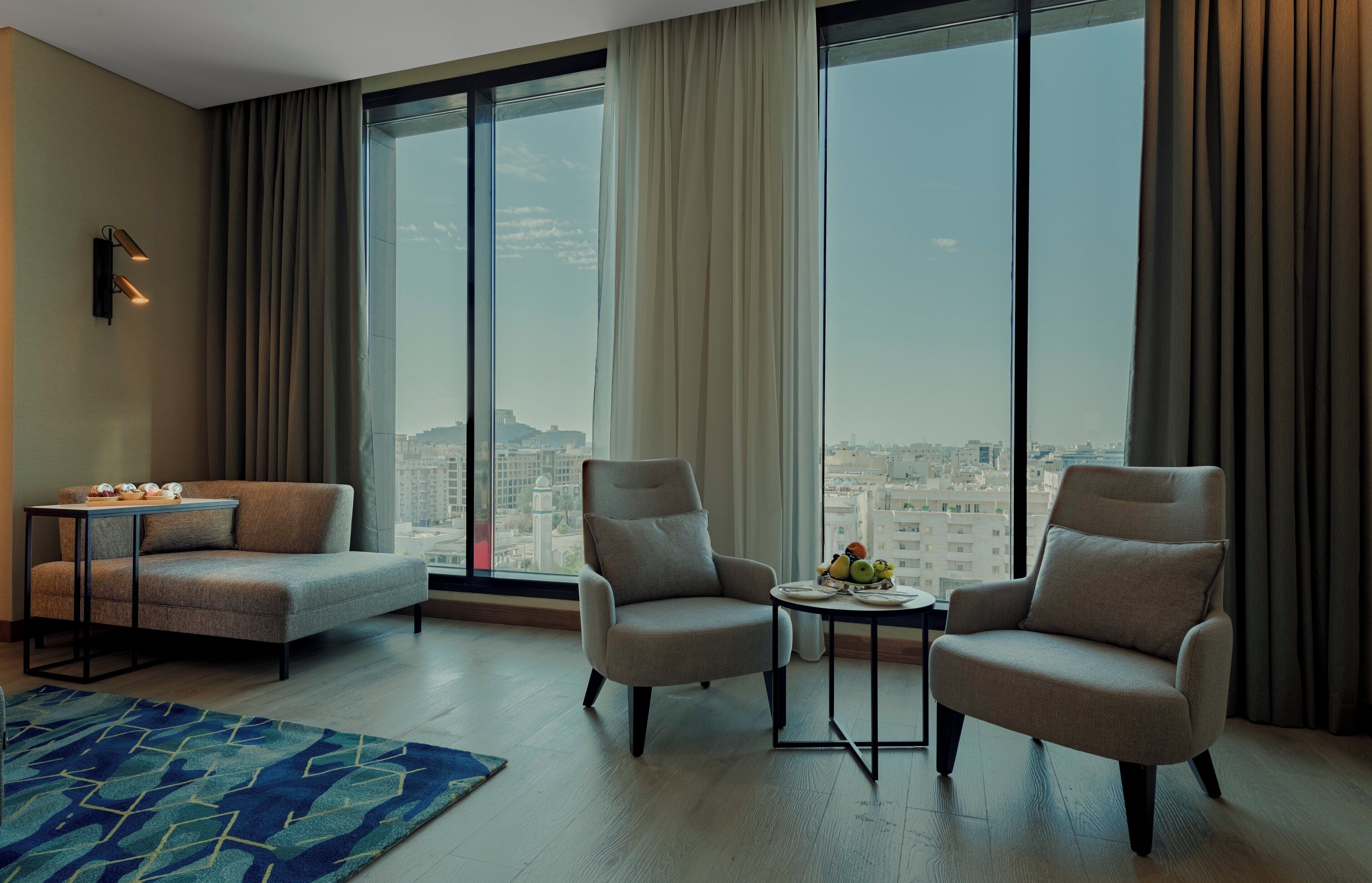 A modern hotel room with a large window, a sofa, two armchairs, a small table with fruit, and a blue patterned rug.