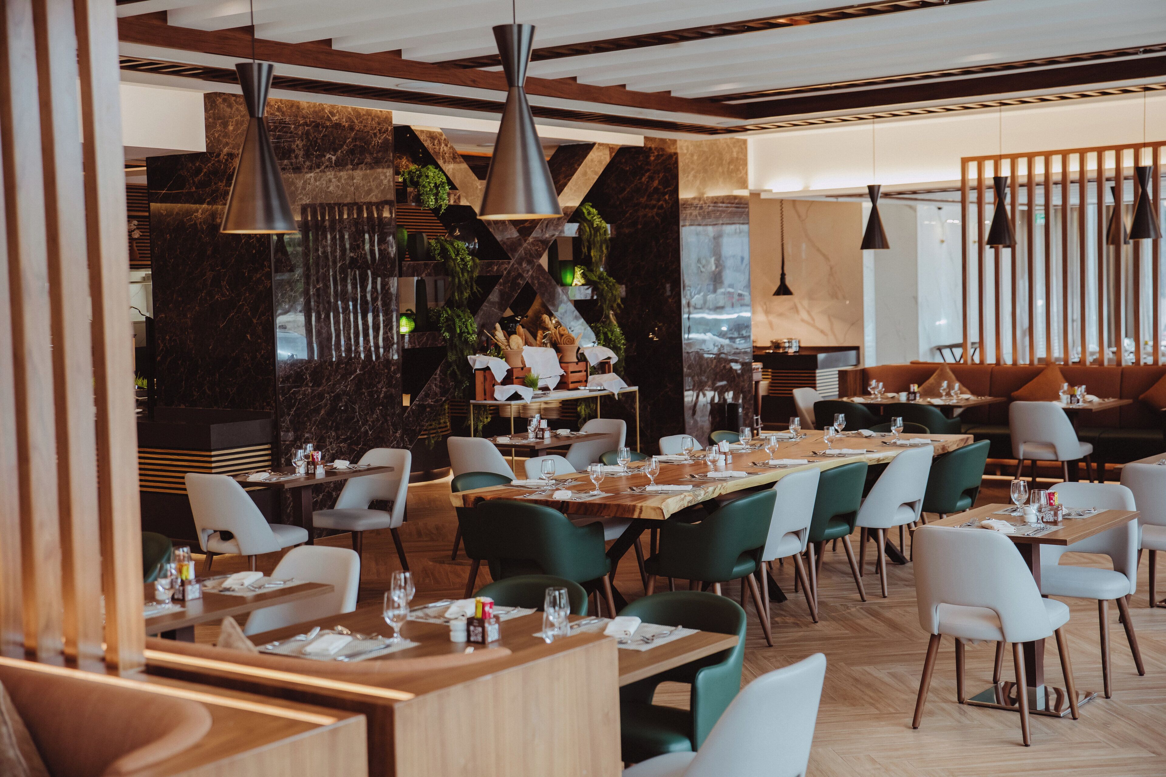 A modern restaurant with wooden tables and green chairs, marble walls, and hanging pendant lights.
