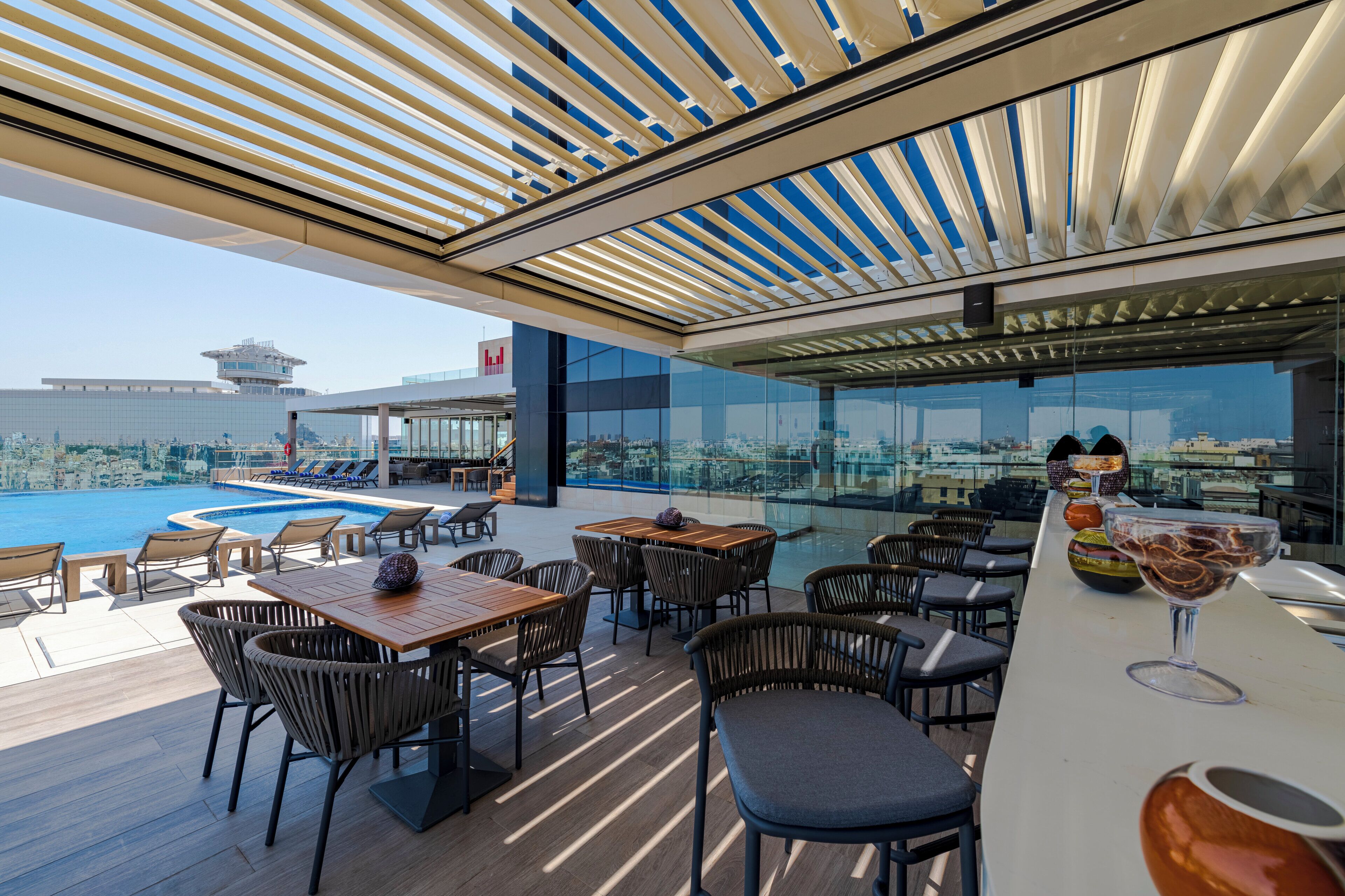 Un bar en la azotea con mesas, sillas, piscina y vistas a la ciudad.