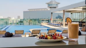A dining table with a plate of fruit and a glass of iced coffee, overlooking a pool and lounge area. - Millennium Al Masar Hotel (Jeddah)
