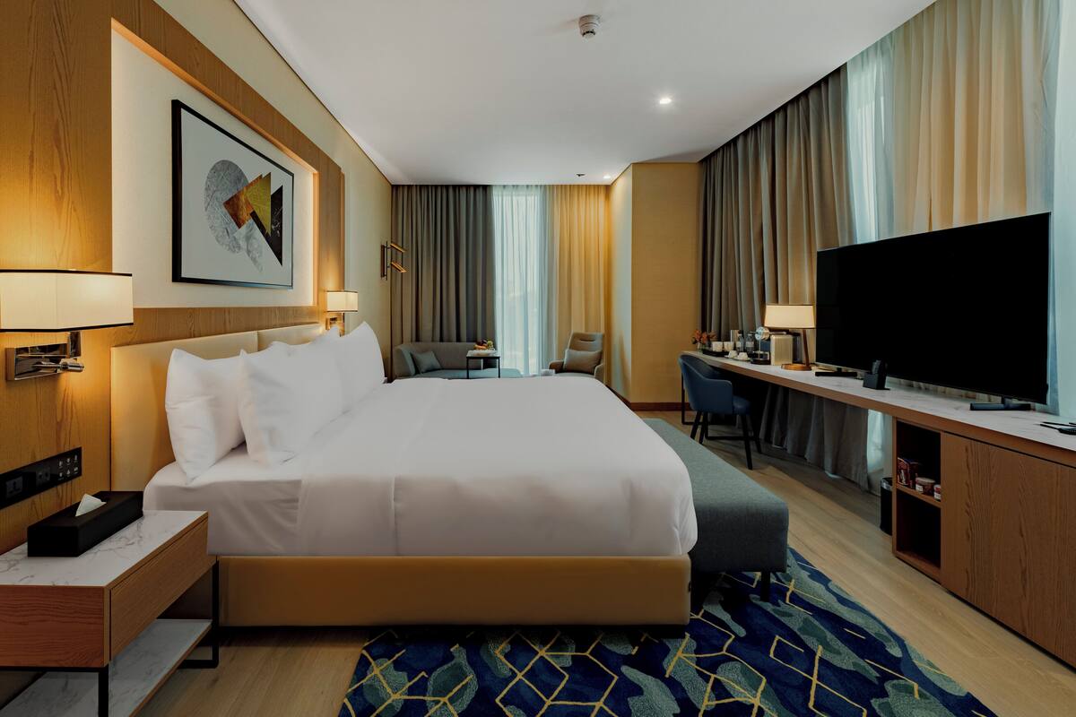 A hotel room with a large bed, a desk with a TV, a chair, and a small table with a fruit bowl.