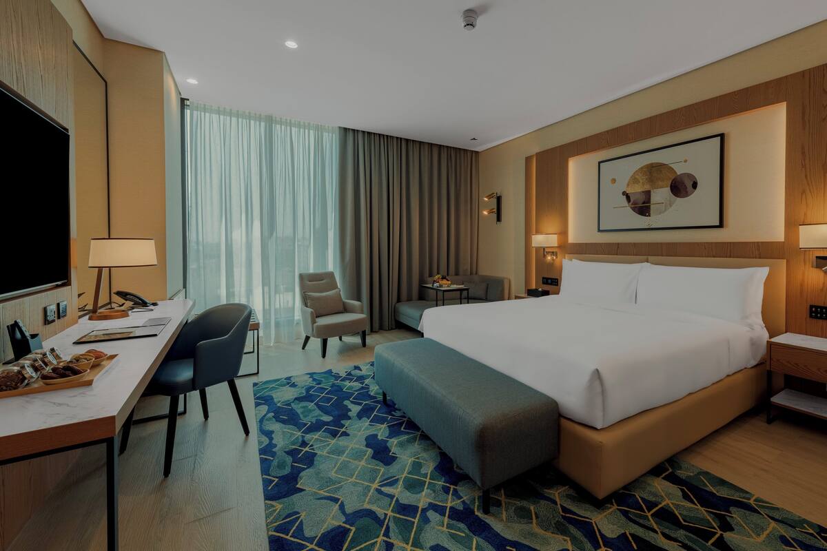 A hotel room with a large bed, a desk with a chair, a small table with a lamp, and a TV.