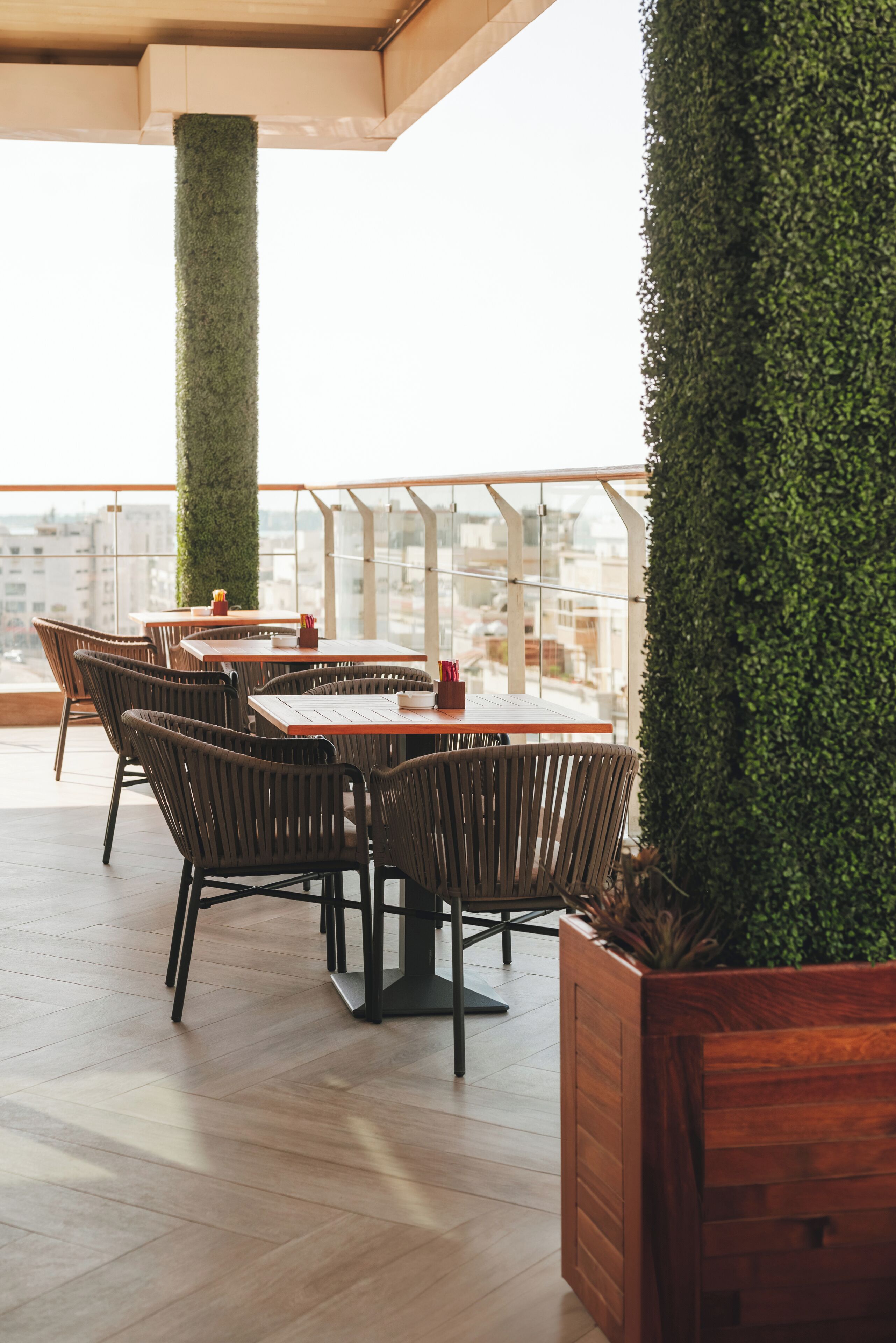 Una terraza en la azotea con mesas y sillas de madera, un seto verde alto y vistas de la ciudad.