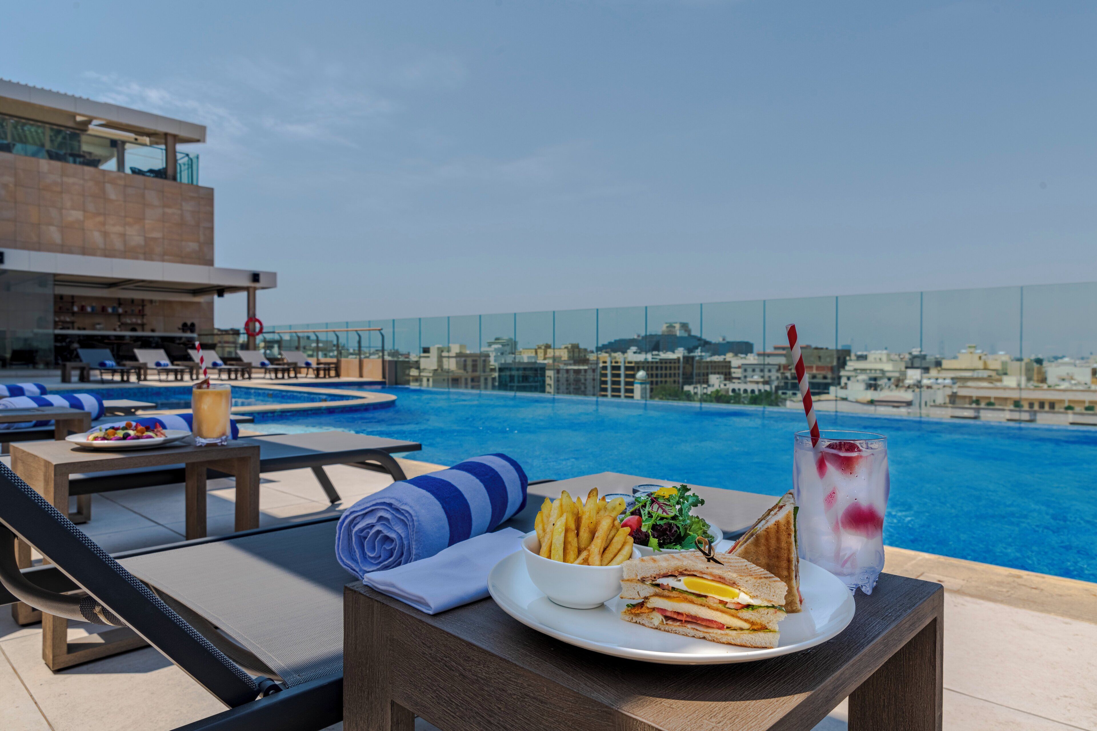 Zona de comedor junto a la piscina con vistas a la ciudad, que incluye una mesa con un sándwich, papas fritas, ensalada y una bebida.