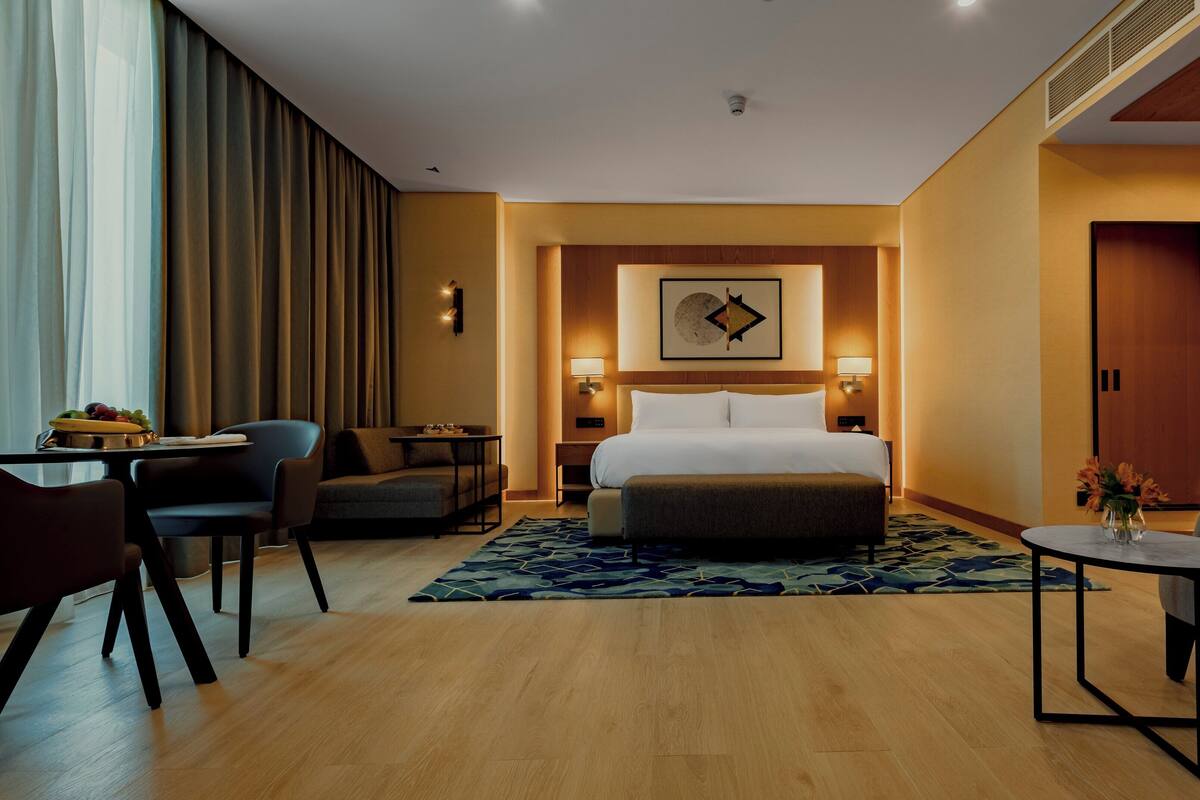 A hotel room with a bed, two armchairs, a small table, a desk, and a dining table with fruit.