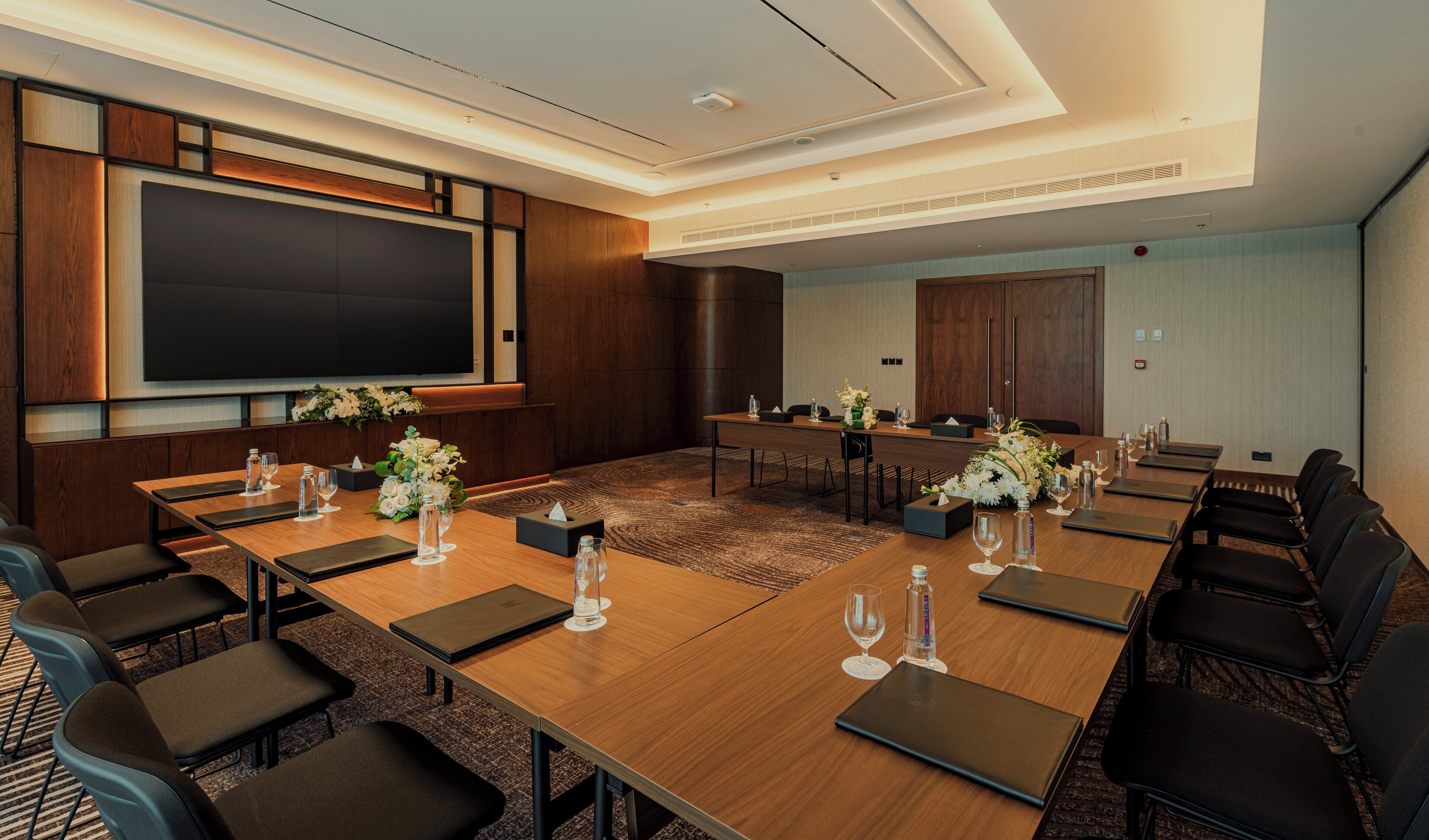 A conference room with a large screen, multiple tables, chairs, and floral centerpieces.