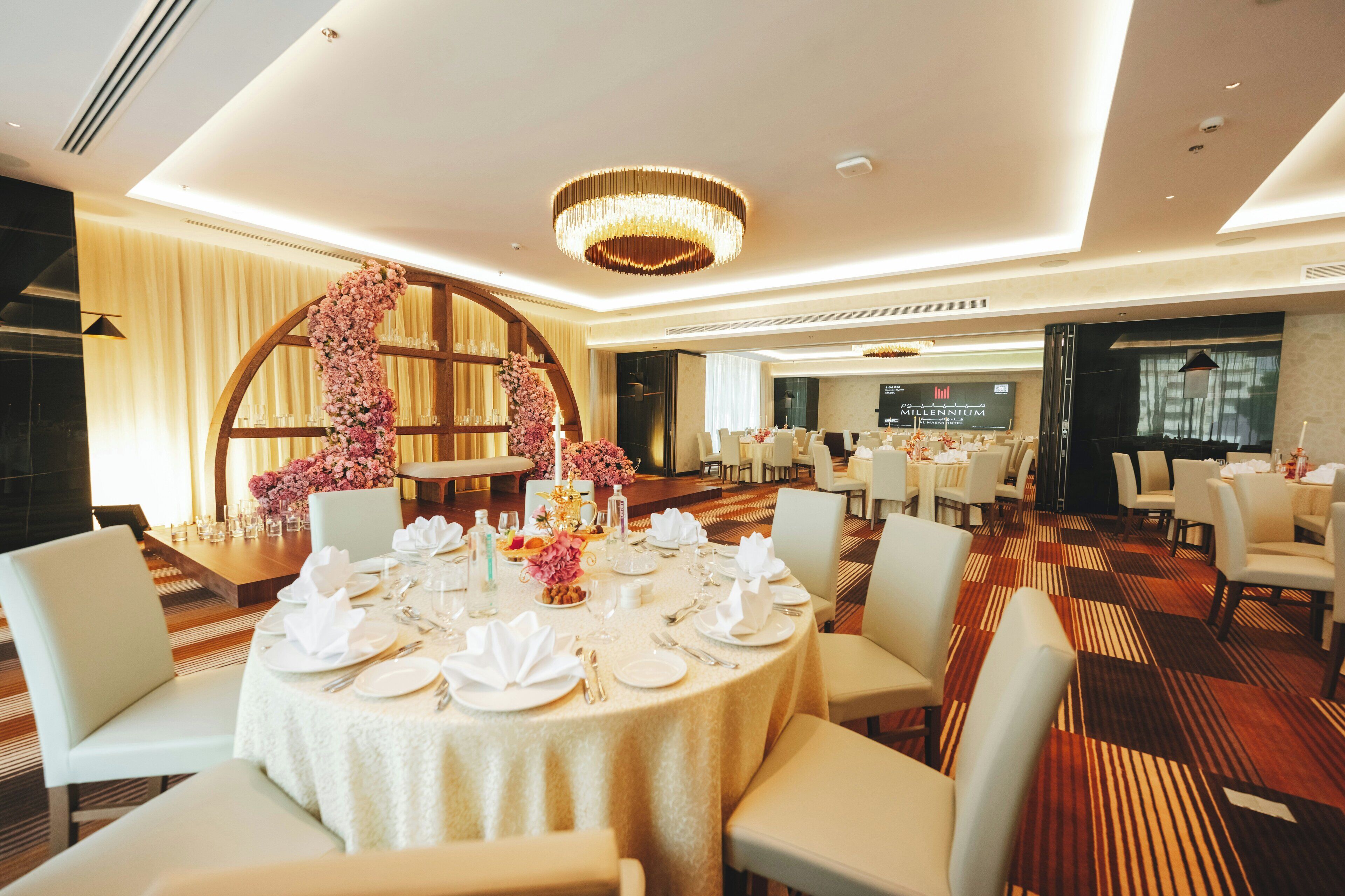 Comedor de hotel con mesas redondas dispuestas para el servicio, una lámpara de araña grande y un arco floral.