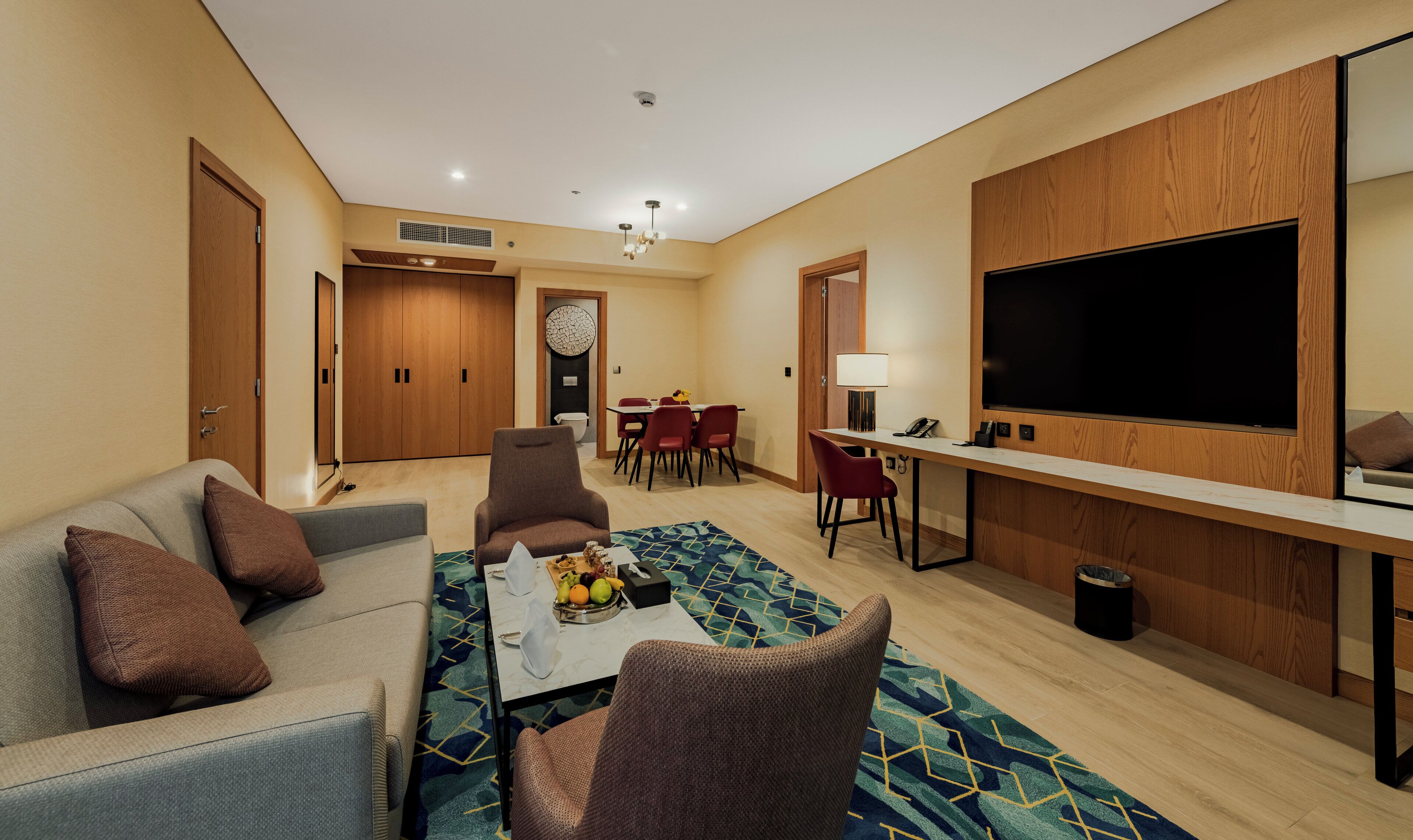 A modern hotel room with a sofa, chairs, a dining table, and a large TV.