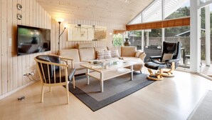 A modern living room with a wooden ceiling, a flat-screen TV mounted on the wall, a beige sofa, a coffee table, and a leather armchair.
