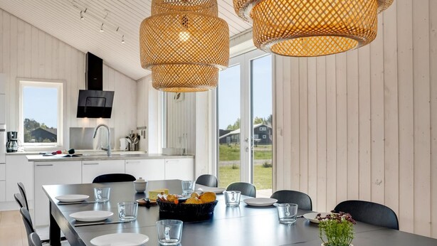 Une cuisine moderne avec des meubles blancs, un grand îlot central et un coin repas doté d’une longue table prévue pour quatre personnes.